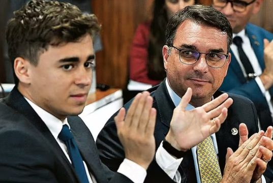 O deputado federal Nikolas Ferreira (PL-MG) e o senador Flávio Bolsonaro (PL-RJ) sentados lado a lado, o primeiro batendo palmas