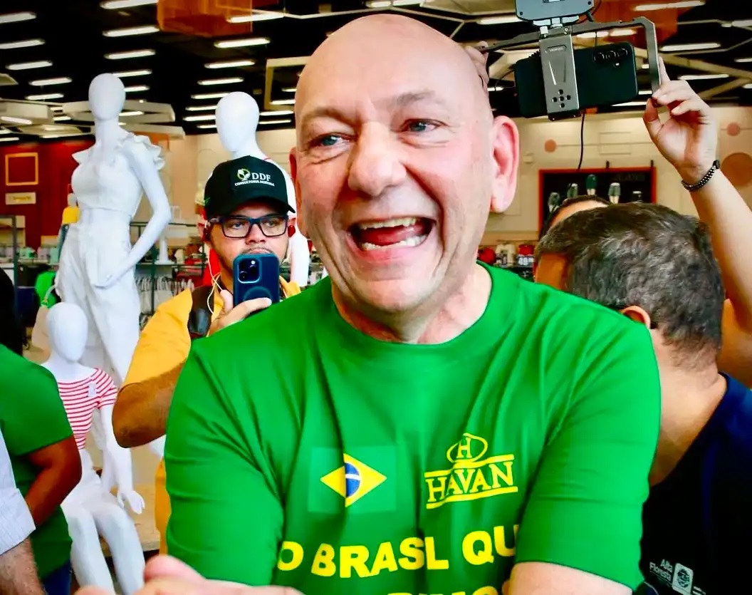 Luciano Hang de braços cruzados, rindo, sem olhar para a câmera, de camiseta verde