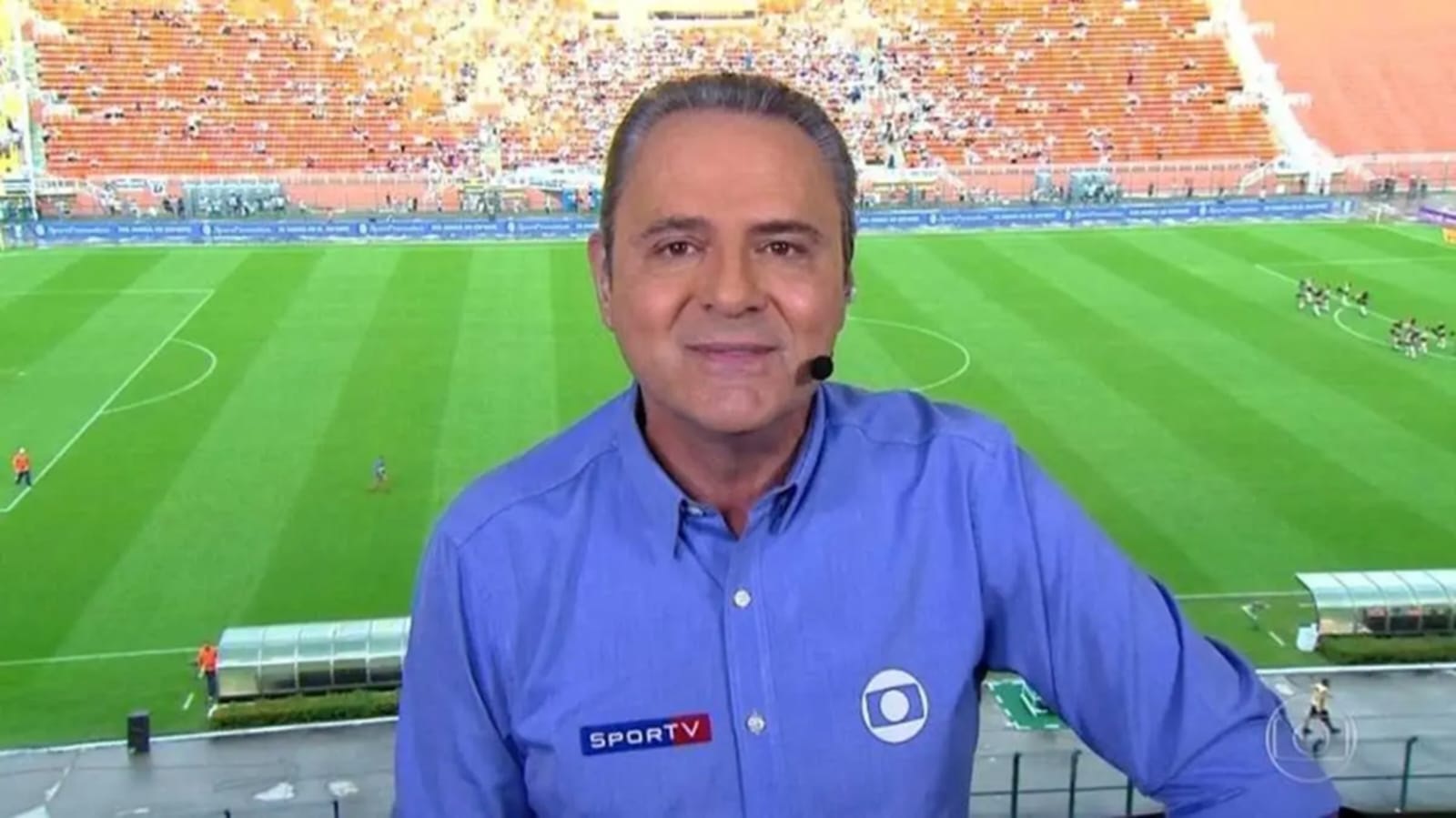 Luis Roberto falando, de camisa social azul, na frente de campo de futebol