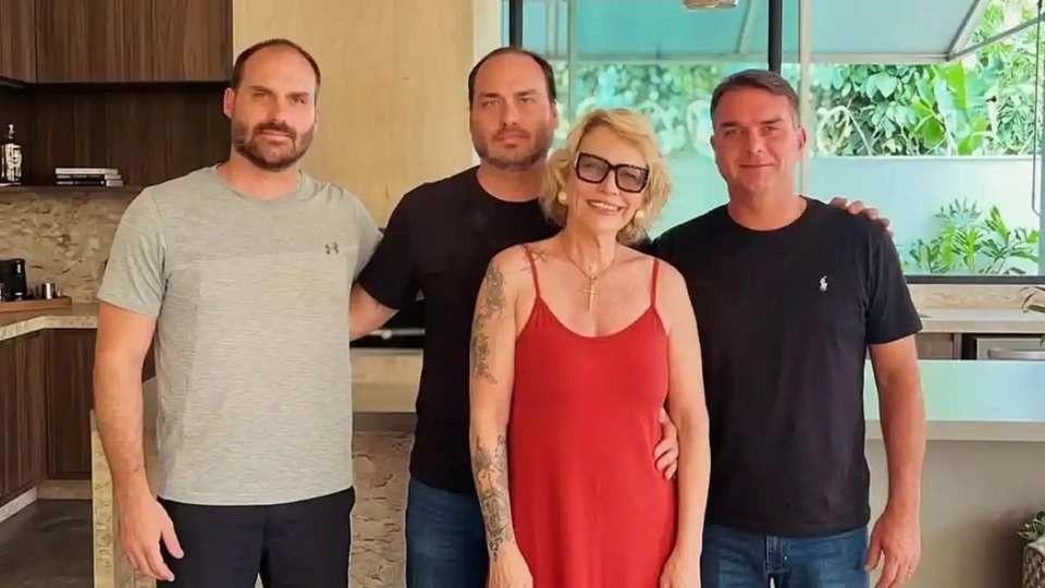 Rogéria Bolsonaro com os filhos Eduardo, Carlos e Flávio, em pé, lado a lado, sorrindo para a câmera
