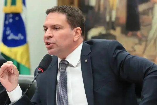 Alexandre Ramagem de perfil, sentado, falando e gesticulando, sério