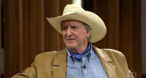 Sérgio Reis de roupa social e chapéu, sentado, falando, olhando para o lado