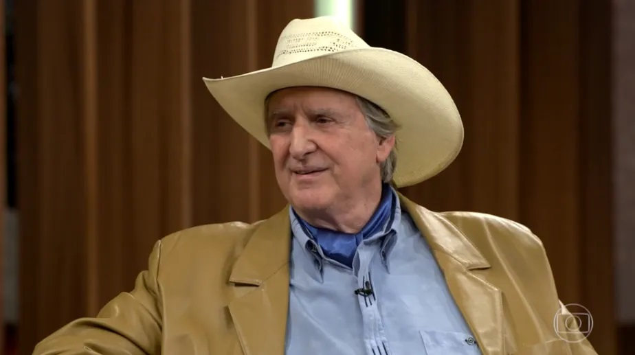 Sérgio Reis de roupa social e chapéu, sentado, falando, olhando para o lado