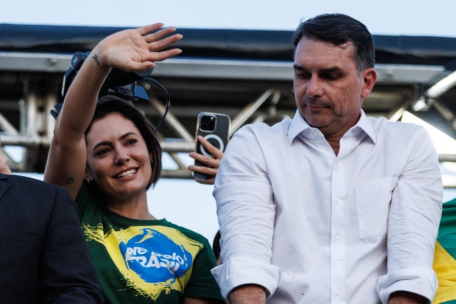 Michelle Bolsonaro acena ao público, enquanto Flávio Bolsonaro apresenta cara de mágoa