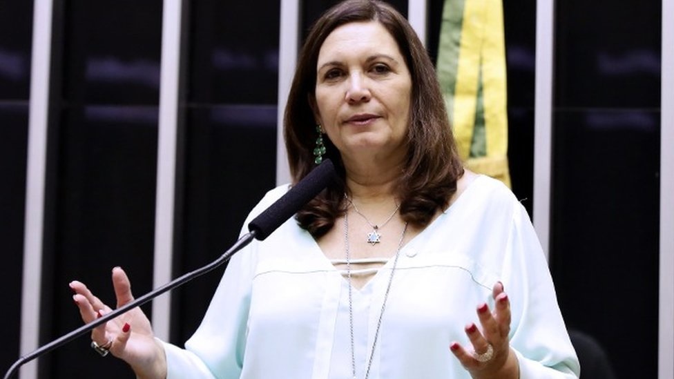 A deputada federal Bia Kicis (PL-DF) de branco, falando em microfone e gesticulando, de cabelo solto