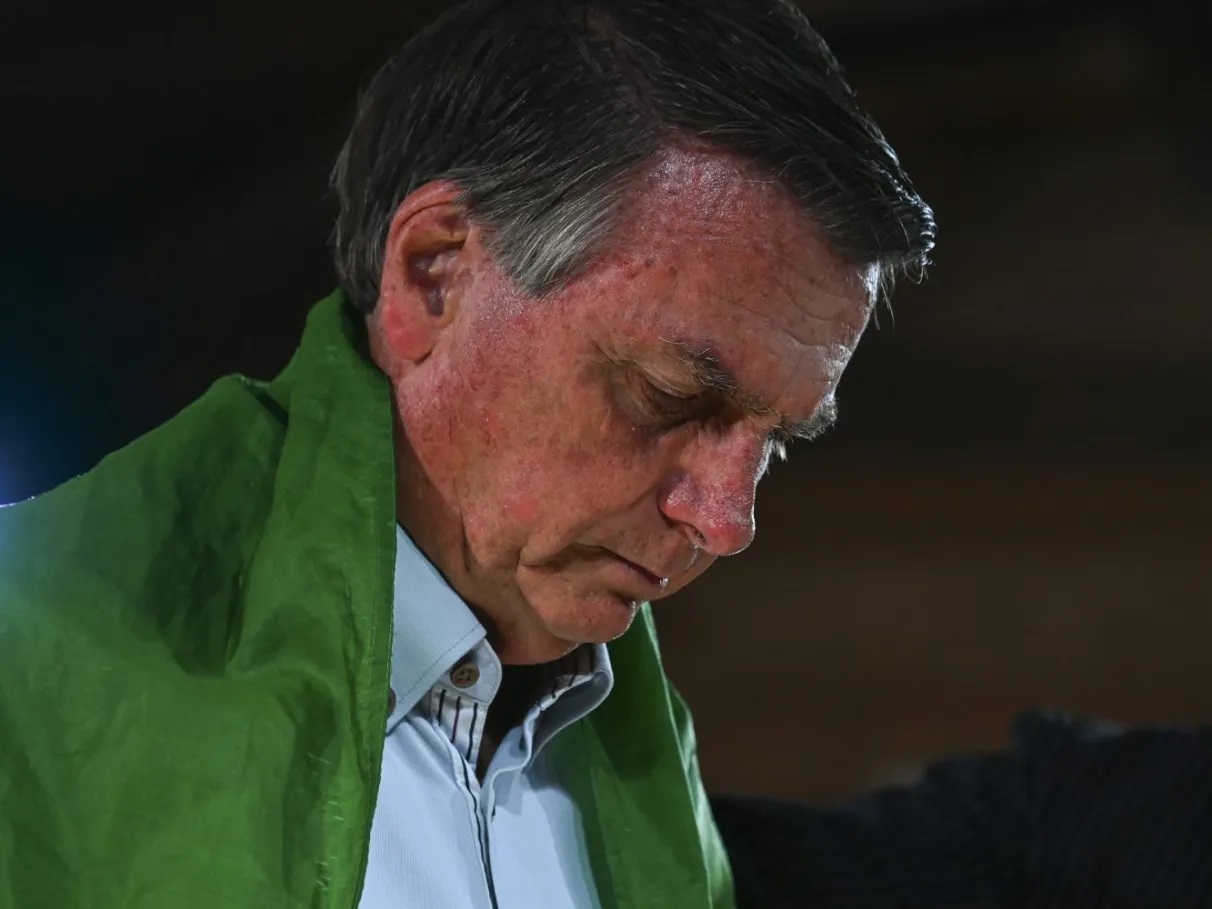 Jair Bolsonaro cabisbaixo, sério, envolto em bandeira do Brasil