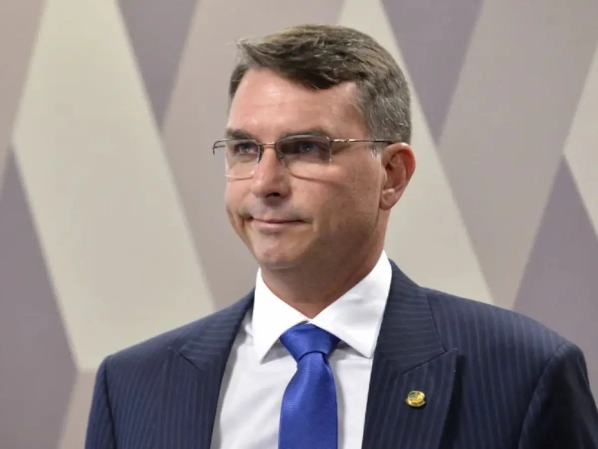 Flávio Bolsonaro com expressão de chateação, roupa social e óculos de grau