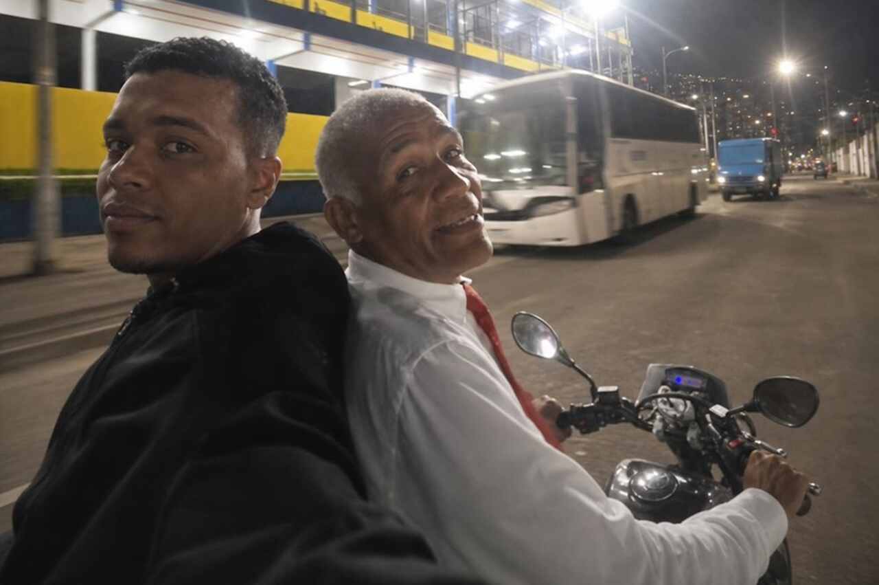 Sargento Isidório em motocicleta ao lado de um jovem em via urbana iluminada