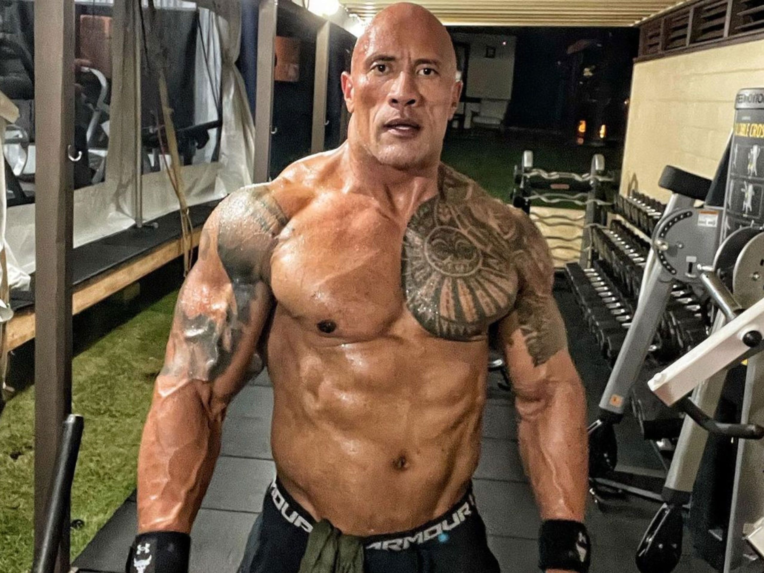 O ator Dwayne Johnson sem camisa, em academia, olhando sério para a câmera