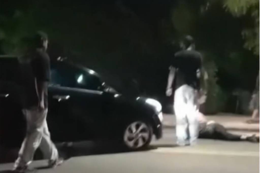 Duas pessoas perto de um carro preto em rua à noite, com uma pessoa caída próxima à calçada