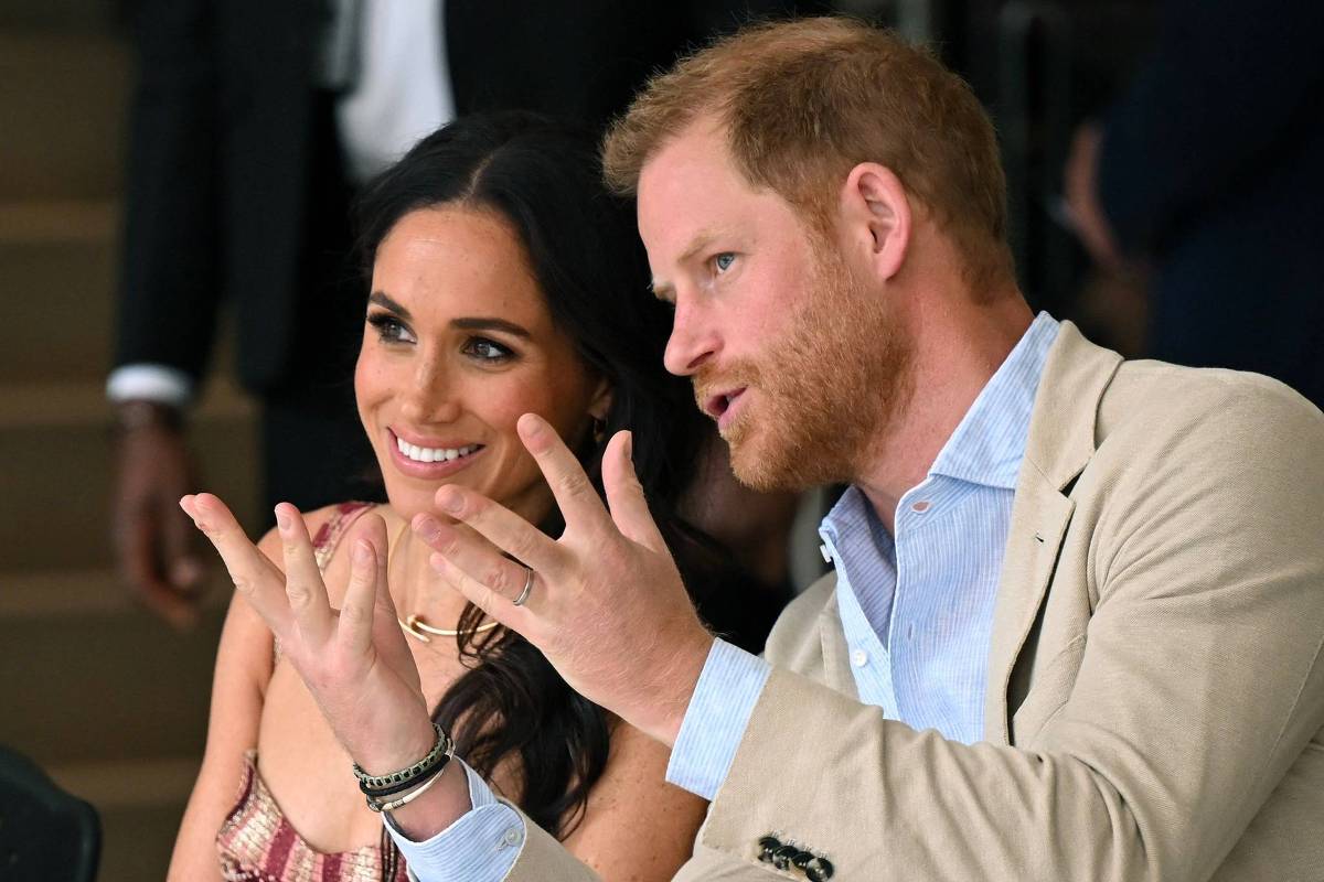 Príncipe Harry fala enquanto Meghan Markle sorri ao seu lado