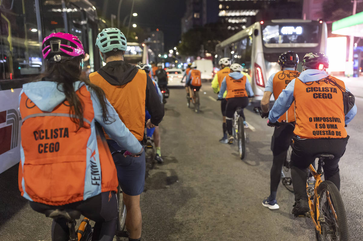 Ciclistas com coletes laranja pedalam à noite em rua de São Paulo