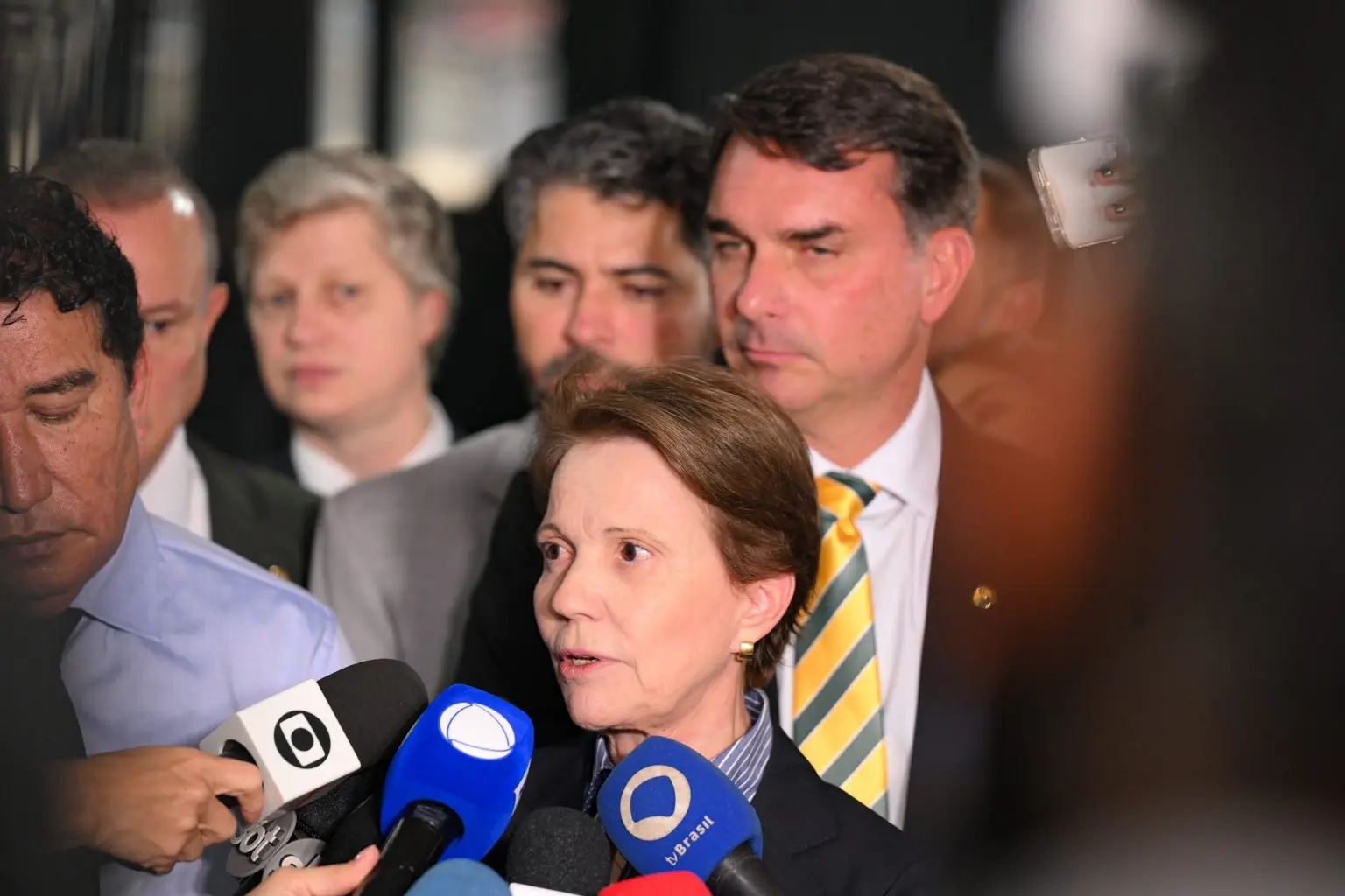 Tereza Cristina concede entrevista com Flávio Bolsonaro ao fundo