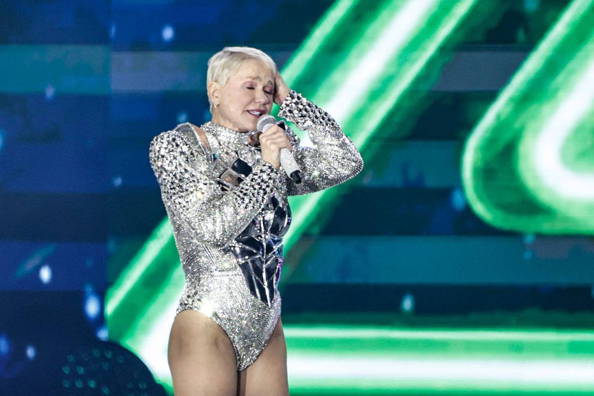 Xuxa canta com body prateado e microfone em palco iluminado