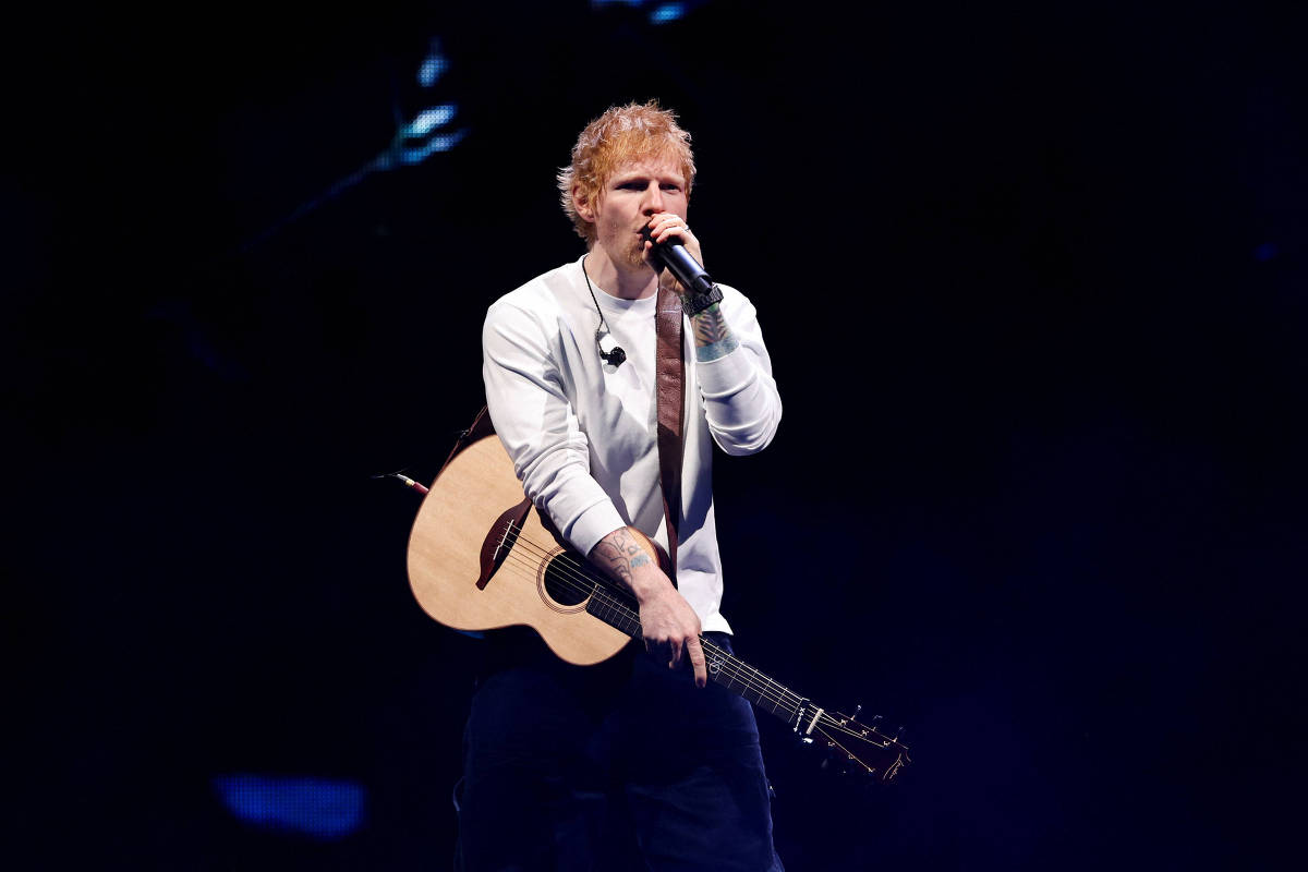 Ed Sheeran canta e toca violão em palco escuro