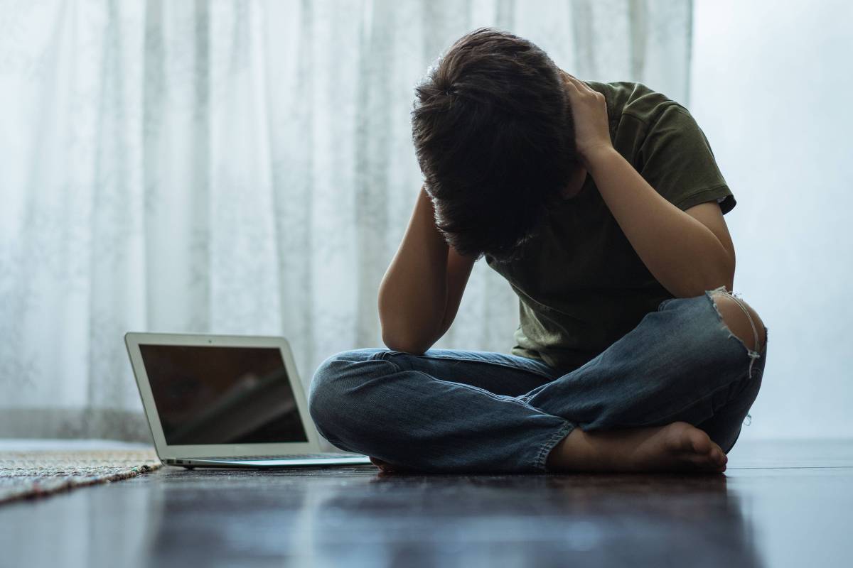 Jovem sentado no chão com as mãos na cabeça ao lado de um laptop