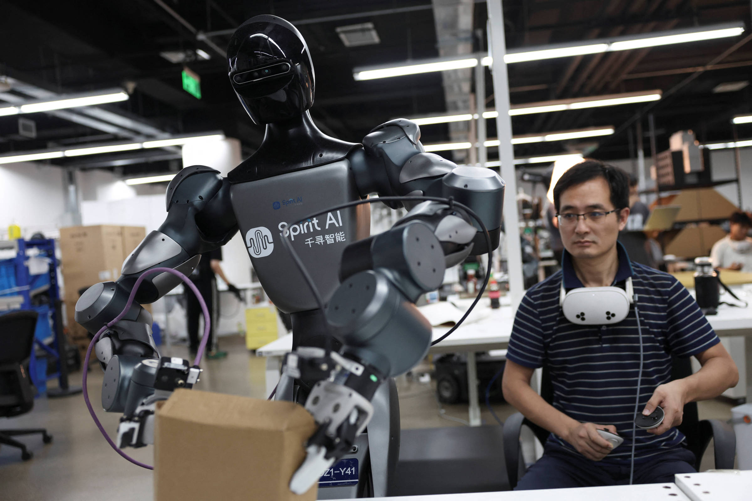 Robô humanoide manipula caixa de papelão enquanto funcionário acompanha o teste