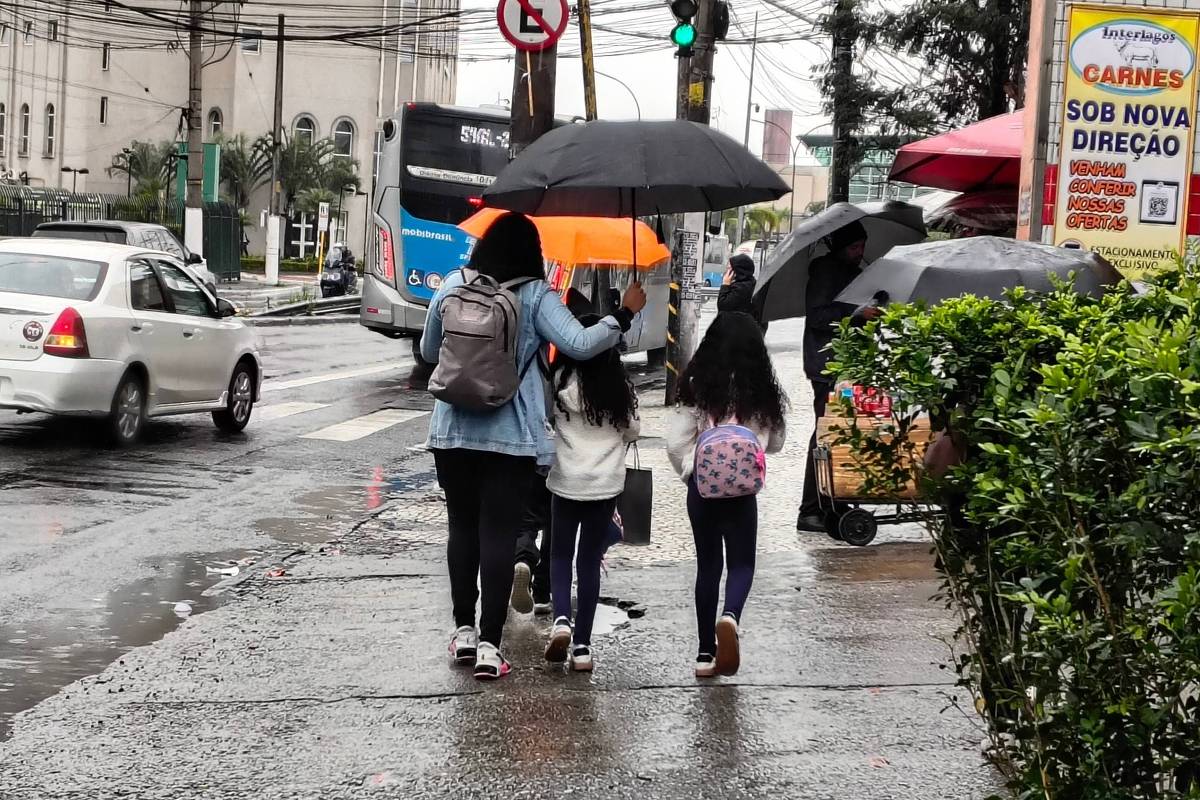 Pedestres caminham com guarda-chuvas em calçada molhada durante chuva em São Paulo