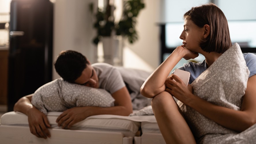 Mulher pensativa deitada ao lado de homem dormindo