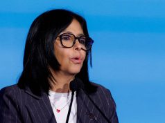 Delcy Rodríguez de óculos e terno, falando ao microfone diante de fundo azul