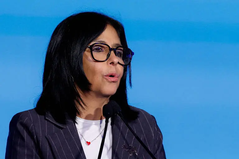 Delcy Rodríguez de óculos e terno, falando ao microfone diante de fundo azul