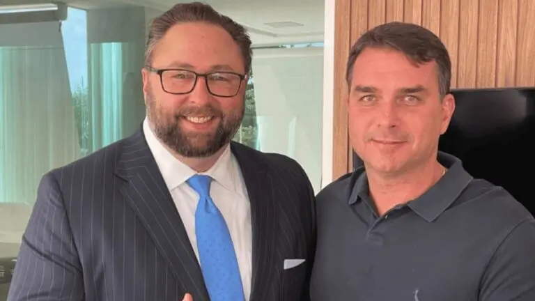 Jason Miller, de terno risca de giz e gravata azul, e Flávio Bolsonaro, de camisa polo escura, posam lado a lado fazendo sinal de positivo