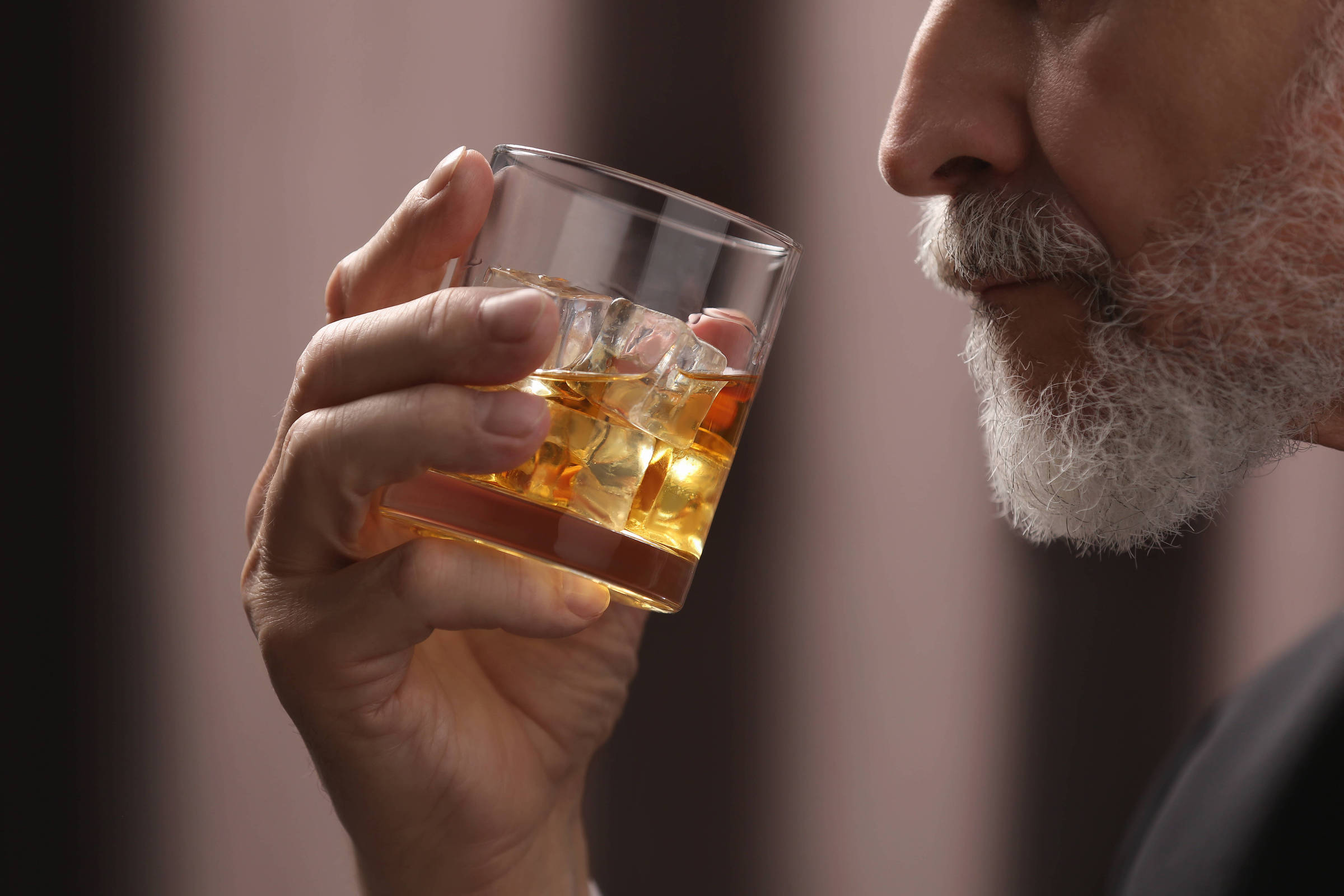 Homem idoso segura copo com bebida âmbar e cubos de gelo