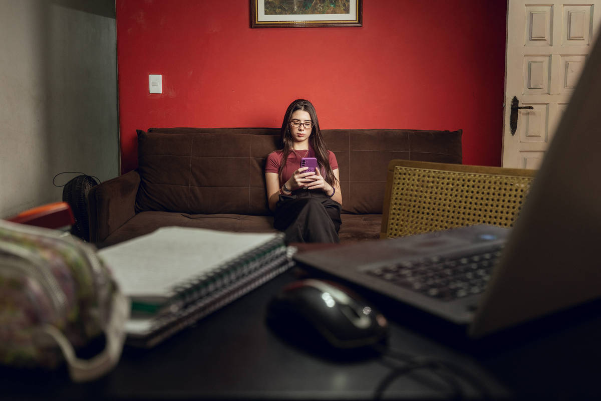 Estudante sentada em sofá olha para celular durante pausa nos estudos