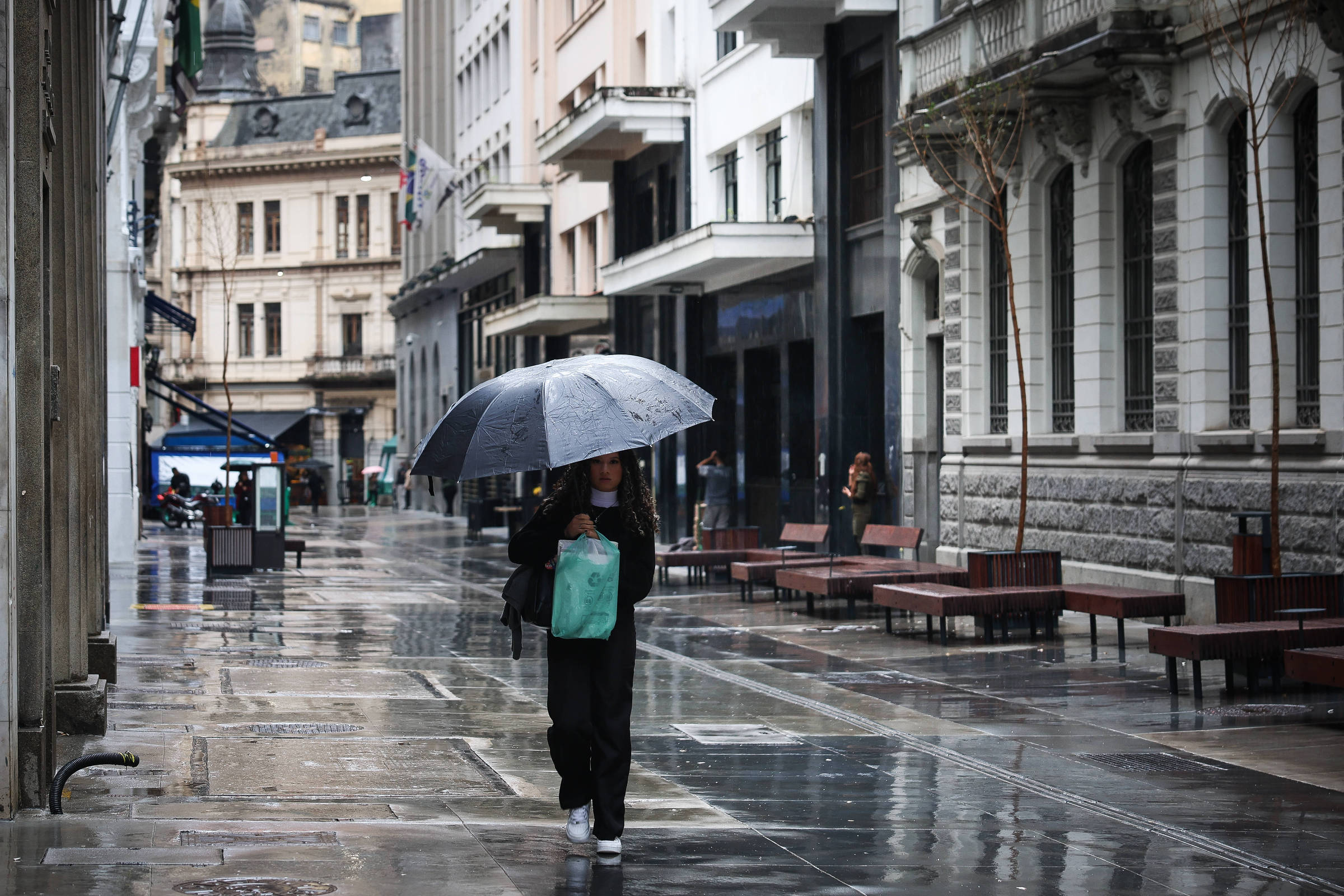 Pedestre com guarda-chuva caminha por rua molhada no centro de São Paulo